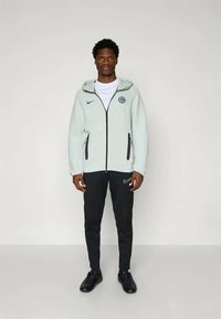 Light gray fleece zip-up hoodie with black accents, logo on chest, paired with black slim-fit pants and black athletic shoes.