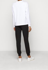 Felpa bianca con collo rotondo, abbinata a pantaloni jogger neri con una striscia dorata laterale. Il modello indossa sneakers bianche.