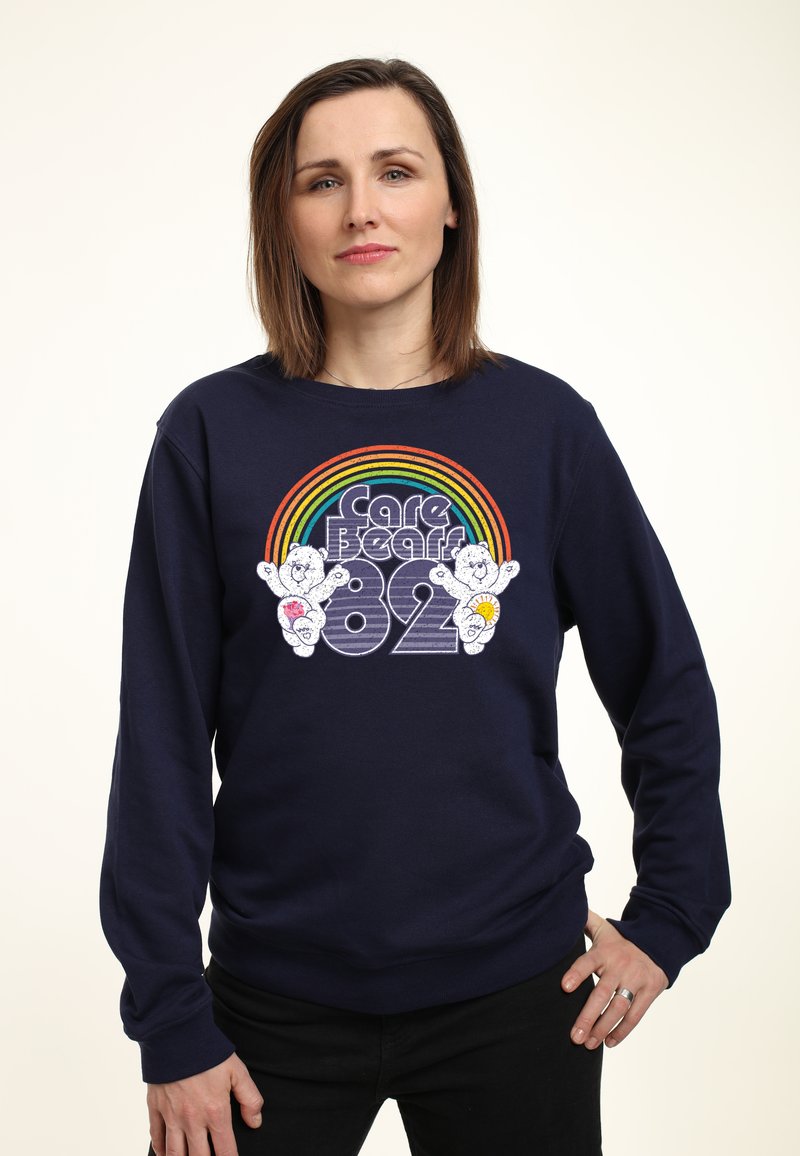 Žena s hnědými vlasy po ramena, oblečená v tmavě modré mikině s motivem dvou Medvídků starostlivosti pod duhou a nápisem "Care Bears 82".