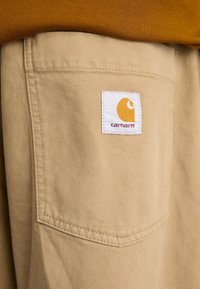 Lähellä oleva kuva beigenvärisistä Carhartt-housujen taskusta, jossa on logo-patch, käytetty ruskean yläosan kanssa.