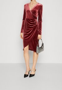 Femme portant une robe portefeuille en velours bordeaux à manches longues, des talons noirs avec des sangles ornées de bijoux, et tenant un sac à main en argent scintillant.