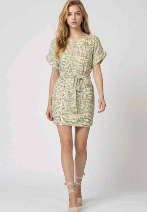 Joven mujer con un vestido corto beige con estampado, cintura anudada y zapatos beige con cordones, de pie frente a un fondo claro y liso.