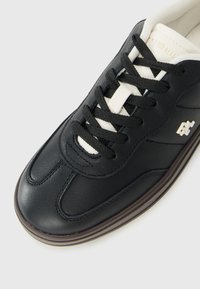 Close-up van een zwart leren sneaker met zwarte veters, een witte tong en een klein wit logo aan de zijkant, tegen een effen witte achtergrond.