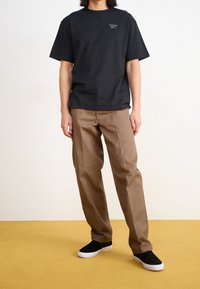 Svart bomullströja med Reebok-logotyp, ihop med ljusbruna byxor och svarta skor med vita sulor, stående på ett gult golv.