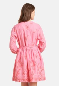 Rotes Kleid mit floralem Stickerei in Koralle. Es hat lange Ärmel, eine taillierte Taille und einen schwingenden Rock. Aus glattem Baumwollstoff.