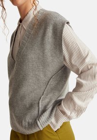 Grau gestrickte Weste mit V-Ausschnitt, strukturiertem Oberflächen, über einem gestreiften Langarmshirt getragen, ergänzt durch senfgelbe Hose.