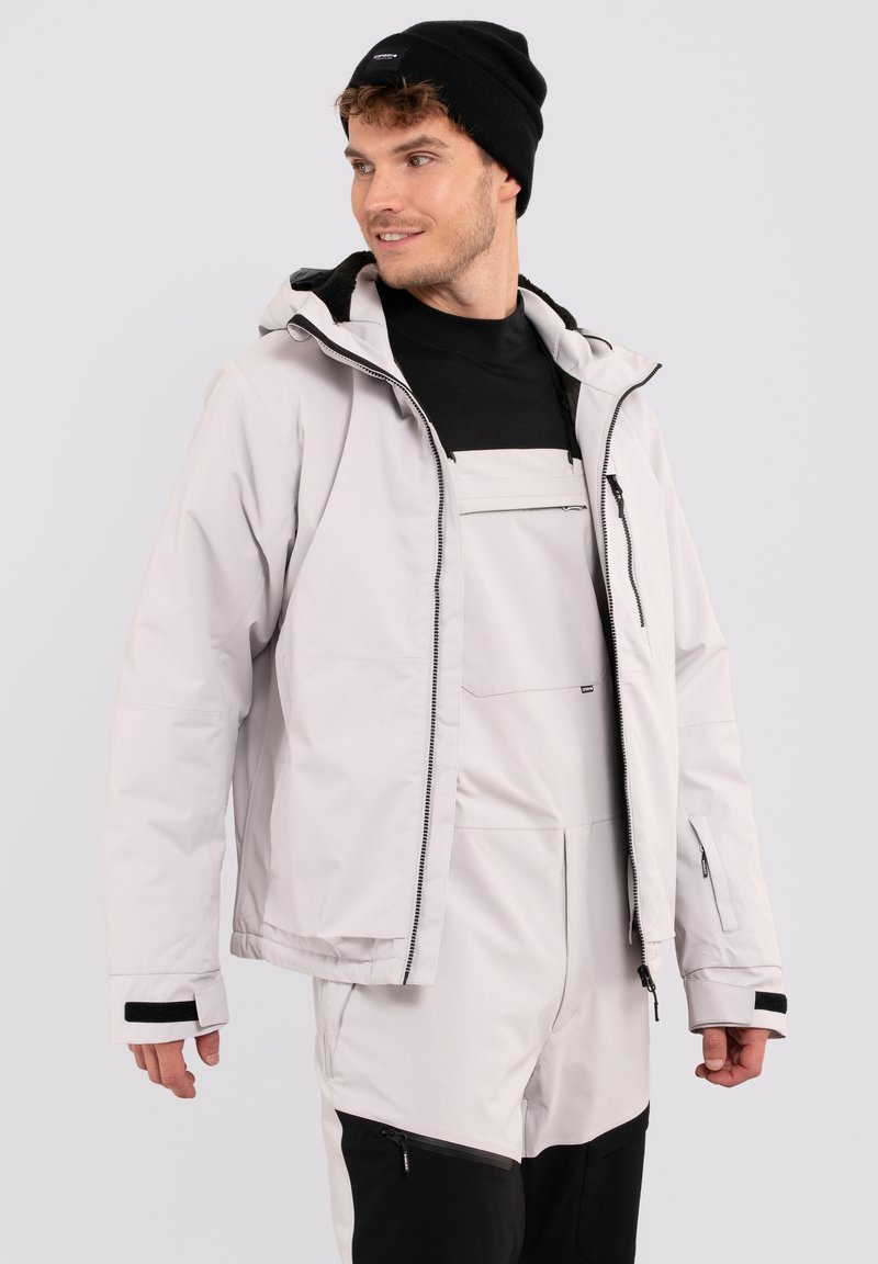 Lichtgrijze skijack met een capuchon, ritsen en verstelbare manchetten, gedragen over een zwart shirt en bijpassende overall. Inclusief zwarte gebreide muts.