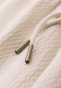 Gros plan d'un tissu blanc texturé avec deux embouts de lacets beiges posés dessus.