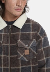 Chaqueta de forro polar a cuadros marrón y gris con un collar de sherpa blanco y un bolsillo en el pecho con un parche de logo y una cremallera metálica.
