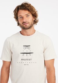Šviesiai smėlinis medvilninis marškinėliai su juodu grafiniu dizainu, kuriame pavaizduotas riedlentė ir banglentė. Yra užrašas "POV PROTEST SPORTSWEAR EST.93".
