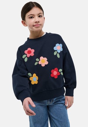 Fille portant un sweat-shirt bleu marine avec des patchs de fleurs colorés et texturés, et un jean bleu, debout devant un fond blanc uni.