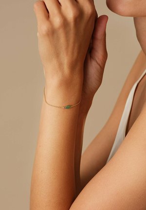 Bracciale in oro con un centro in pietra verde. Il bracciale presenta un design delicato, posato su un polso con pelle liscia.