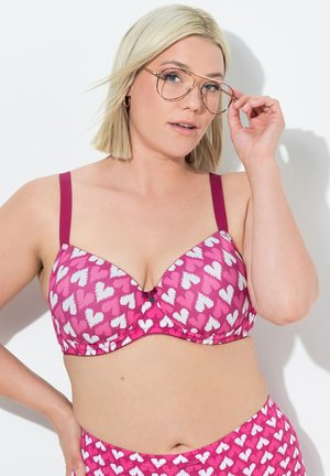 Roze beha met een patroon van witte hartjes, verstelbare kastanjebruine bandjes en een zwarte decoratieve rand. Gladde stof met een lichte glans.