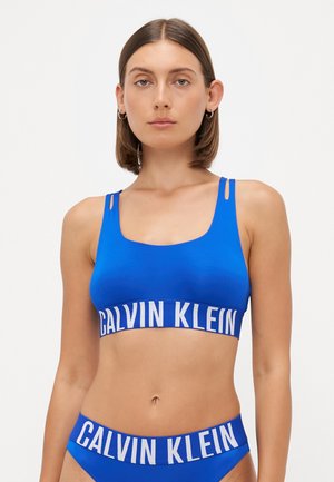Blauwe sportbh met een brede witte Calvin Klein-logo band, voorzien van verstelbare schouderbanden en een gladde textuur. Model draagt bijpassende slip.