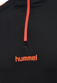 Svart sportstopp med hög krage, med en fjärdedels dragkedja och ljusa orange detaljer. "Hummel"-logotyp i orange på bröstet.