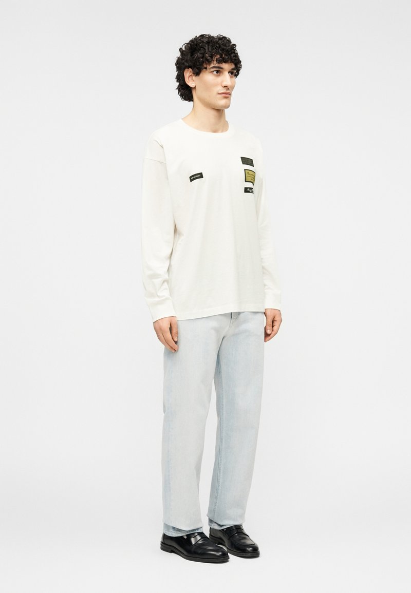 Chemise crème à manches longues avec plusieurs patchs en tissu noir et vert, associée à un jean bleu clair et des chaussures noires, posant devant un fond uni.