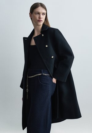 Kobieta w czarnym dwurzędowym płaszczu na czarnej bluzce i granatowych teksturowanych spodniach z paskiem na złoty łańcuch, stojąca na jednolitym tle.