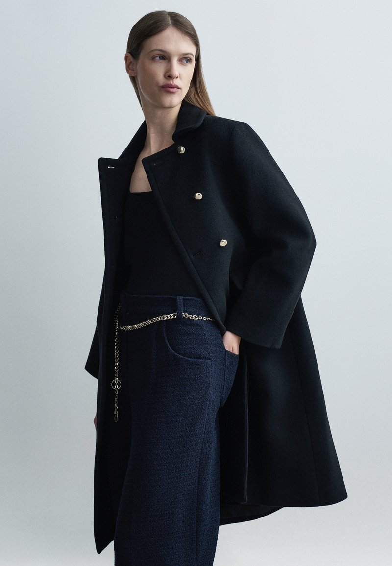 Γυναίκα που φοράει μαύρο διπλό παλτό πάνω από μαύρη μπλούζα και ναυτικό υφασμάτινο παντελόνι με χρυσή αλυσίδα στη μέση, στέκεται μπροστά σε απλό φόντο.