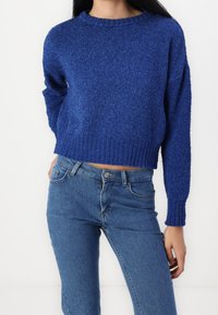 Pull court bleu avec maille texturée, encolure ronde et poignets côtelés, assorti à un jean en denim avec fermeture à boutons.