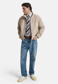 Tan bomber jacket, white shirt, navy striped tie, light blue jeans, and cream sneakers. Model stands with hands in pockets against a white background.