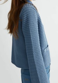 Rakenteellinen sininen lyhyt neuletakki kudotulla kuviolla, jossa on napitus ja rento istuvuus. Pehmeä kangas muodostaa kontrastin sileiden housujen kanssa.