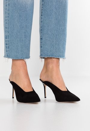 Czarne zamszowe mule o spiczastym nosku i V-kształtnym wycięciu. Oferują wysoką szpilkę i otwarte zapięcie z tyłu, zestawione z krótkimi niebieskimi dżinsami.