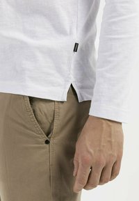 Weißes Longsleeve-Shirt aus strukturiertem Stoff; mit Seitenschlitzen und einem schwarzen Etikett. Kombiniert mit hellbraunen Hosen mit einer bündigen Tasche.