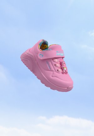 Roze kindersneaker met Care Bears-decoratie, klittenbandsluiting en kleurrijke voering zwevend tegen een blauwe luchtachtergrond.