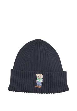 Gorro de punto azul oscuro con borde doblado, con un pequeño oso de peluche bordado que lleva pantalones a rayas y una bufanda verde.