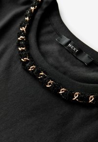 T-shirt noir avec un col texturé orné d'accents noirs et dorés rosés. L'étiquette indique "NEXT". Tissu à texture lisse.
