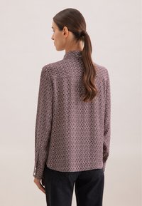 Blouse à manches longues en tissu bordeaux foncé avec un motif géométrique blanc, dotée d'un col à boutons et d'une coupe décontractée à l'arrière.