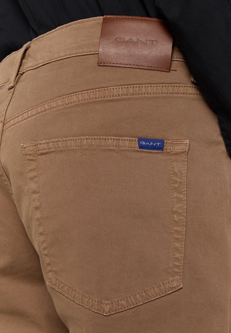 Pantalon en coton marron avec une étiquette en cuir portant le logo "GANT", coutures visibles et une petite étiquette de marque bleue sur la poche arrière.