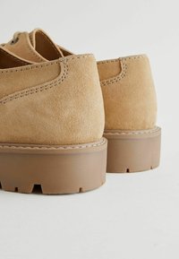 Zapatos de ante color arena con una superficie texturizada, que cuentan con una suela de goma marrón gruesa y costuras visibles a lo largo de los bordes.