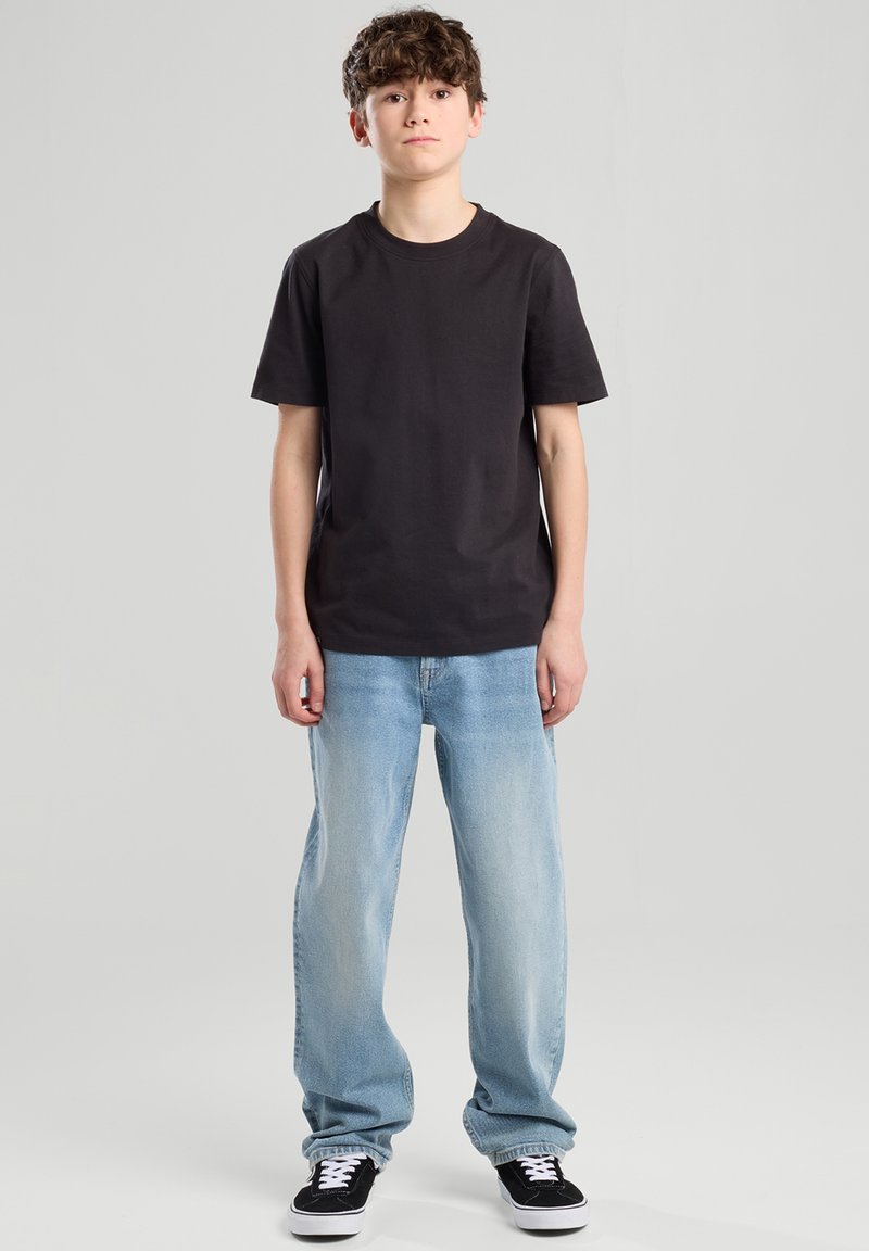 Teen boy standing facing forward wearing black t-shirt, light blue loose jeans, and black sneakers with white laces against plain background.