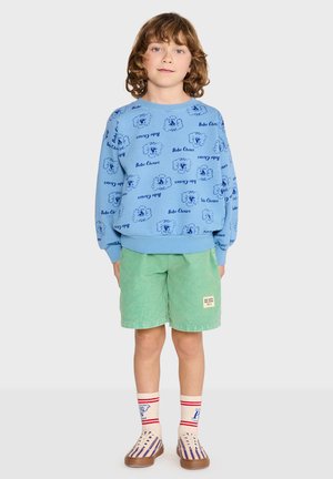Bambino con capelli ricci che indossa una felpa blu stampata, pantaloncini verdi, calzini bianchi con strisce rosse e scarpe slip-on a righe, in piedi su uno sfondo semplice.