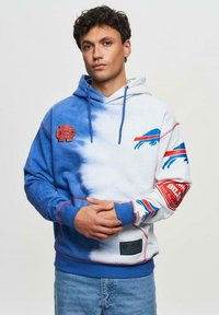 Huppari sinivalkoisessa tie-dye-tyylissä, punaisilla ja sinisillä yksityiskohdilla, jossa joukkueen logot rinnassa ja hihassa. Pehmeä kangas, rento istuvuus.