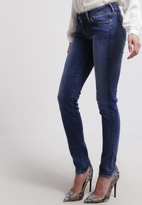 Jeans de mezclilla oscuros ajustados con cinco bolsillos, pasadores para cinturón y una textura suave, combinados con zapatos de tacón alto con un diseño estampado.