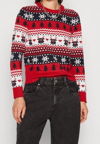 Pull en tricot rouge avec un motif festif de rennes, flocons de neige, cœurs et arbres, doté d'un col et de poignets côtelés. Associé à un pantalon noir.