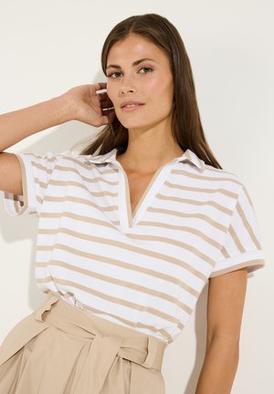 Frau mit langen braunen Haaren, die ein weiß-beiges gestreiftes Kurzarmshirt und beige hoch taillierte Hose mit Gürtel trägt, posiert mit der Hand in der Nähe der Haare.