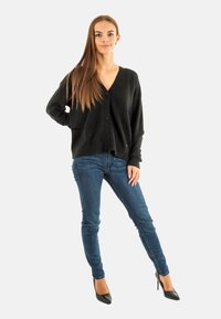 Cárdigan negro de punto con escote en V, mangas largas y botones en el frente, combinado con jeans ajustados azul oscuro y tacones altos negros.