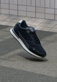 Schwarze Sportschuhe mit einem glatten Lederobermaterial, grauen Akzenten, runden Schnürsenkeln und einer weißen Schaumsohle. Erhaben auf einer strukturierten Oberfläche.