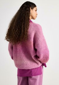 Nadměrný růžový pletený svetr s přechodem barev, který má měkkou texturu a spadlý střih ramen, doplněný fialovým topem pod ním.