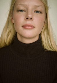 Temno rjav rebrast pulover s turtle neck, prilagojena zasnova, vidna tekstura. Oseba ima svetlo blond lase in naraven make-up, ter modre oči.