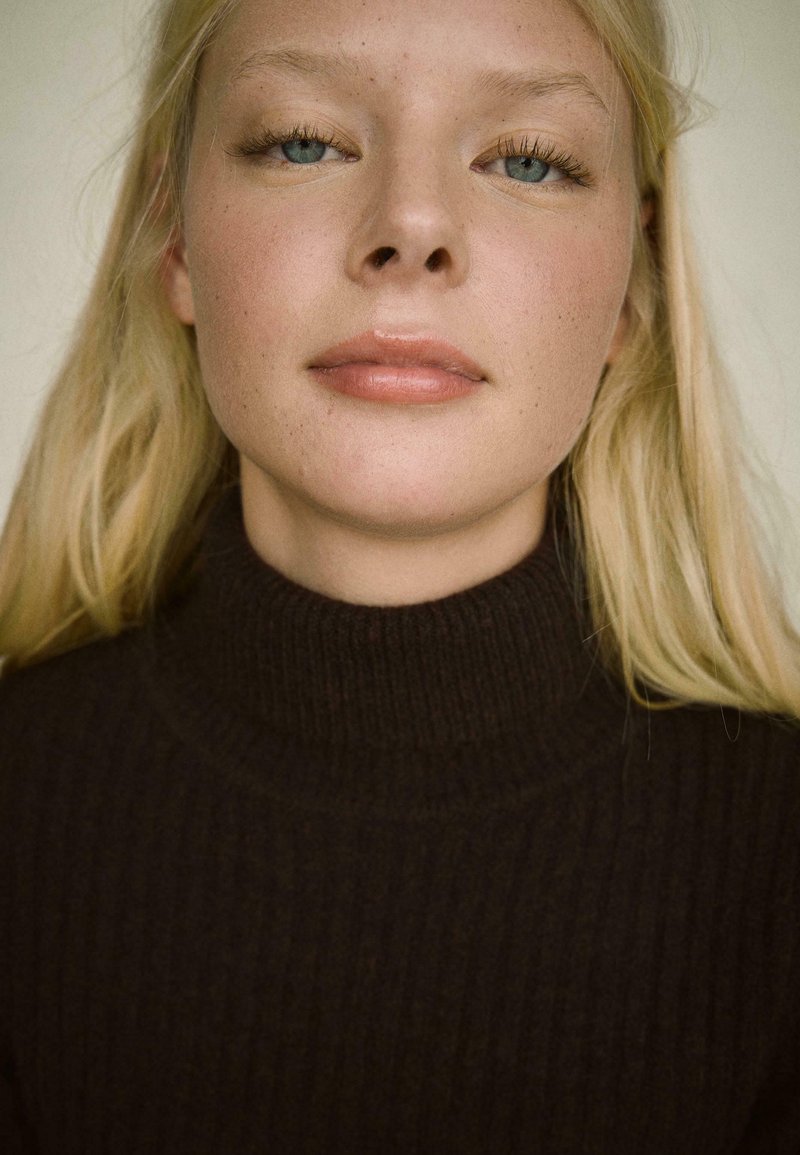 Temno rjav rebrast pulover s turtle neck, prilagojena zasnova, vidna tekstura. Oseba ima svetlo blond lase in naraven make-up, ter modre oči.
