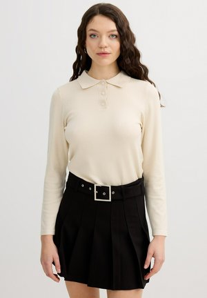 Joven mujer con cabello largo, rizado y castaño, que lleva una camiseta crema de manga larga y una falda negra plisada con un cinturón de hebilla cuadrada, de pie sobre un fondo blanco.