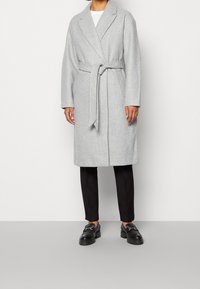 Grå fleece frakke med bånd i taljen, store revers og lange ærmer. Kombineret med sorte slanke bukser og kraftige sorte loafers.