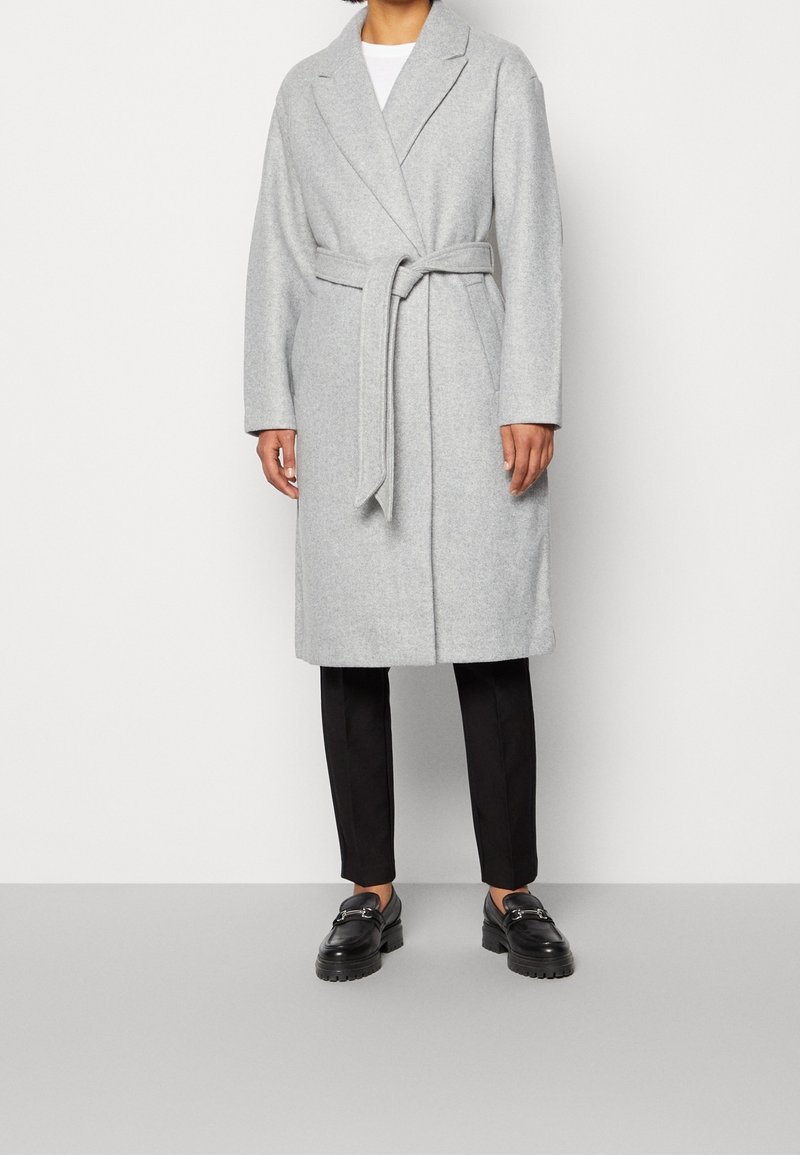 Grå fleece frakke med bånd i taljen, store revers og lange ærmer. Kombineret med sorte slanke bukser og kraftige sorte loafers.