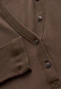 Bruine geribde cardigan met een V-hals, twee knopen en een voorkantzak. De stof heeft een gestructureerde afwerking met subtiele verticale lijnen.