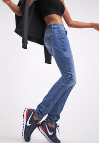 Pantalones vaqueros de mezclilla azul con un corte ajustado, detalles descoloridos y un ligero desgaste. Combinados con zapatillas marinas que tienen detalles en blanco y rojo.