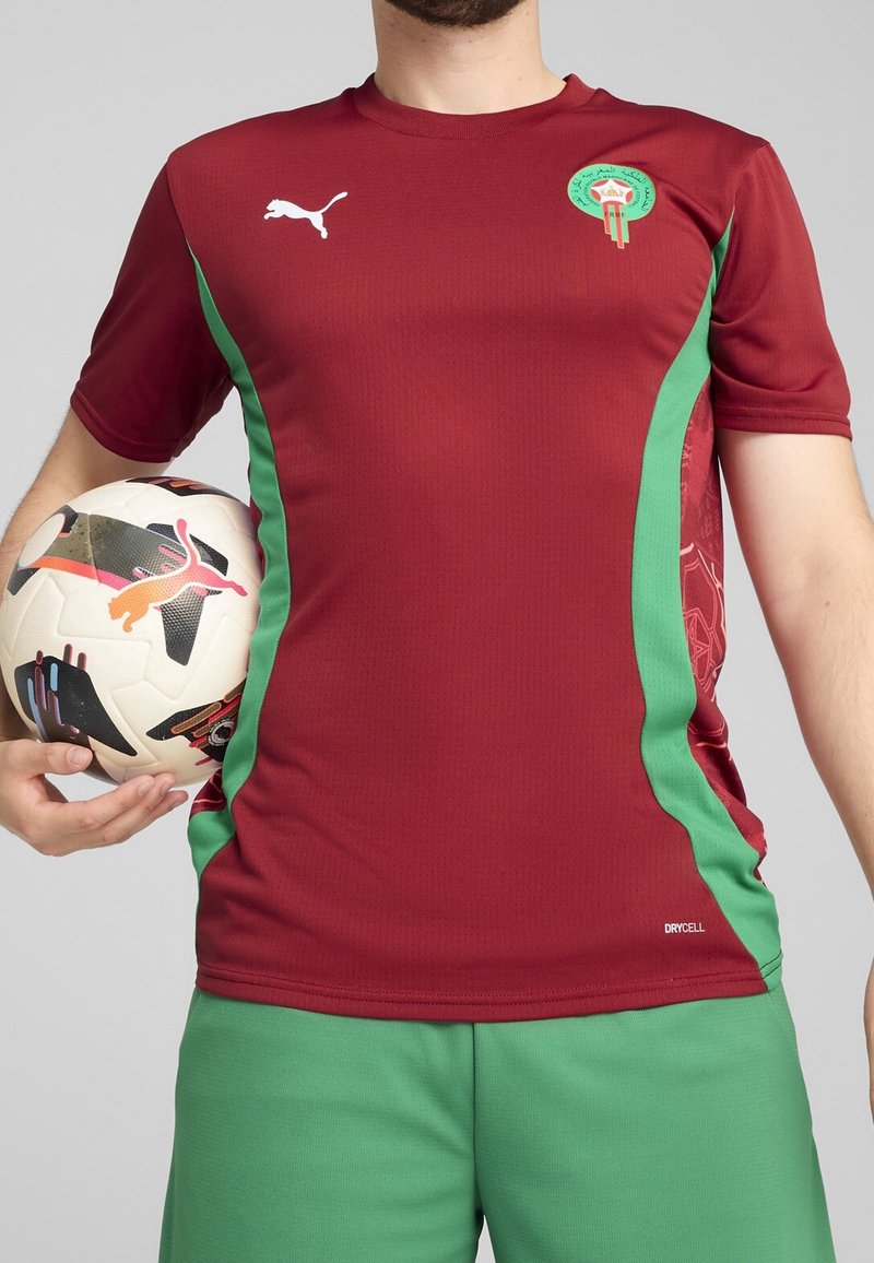 Punainen ja vihreä lyhythihainen jalkapallo-asu, jossa on logo vasemmassa rinnassa ja teksturoitu yksityiskohta sivussa, yhdistettynä vihreisiin shortsit.